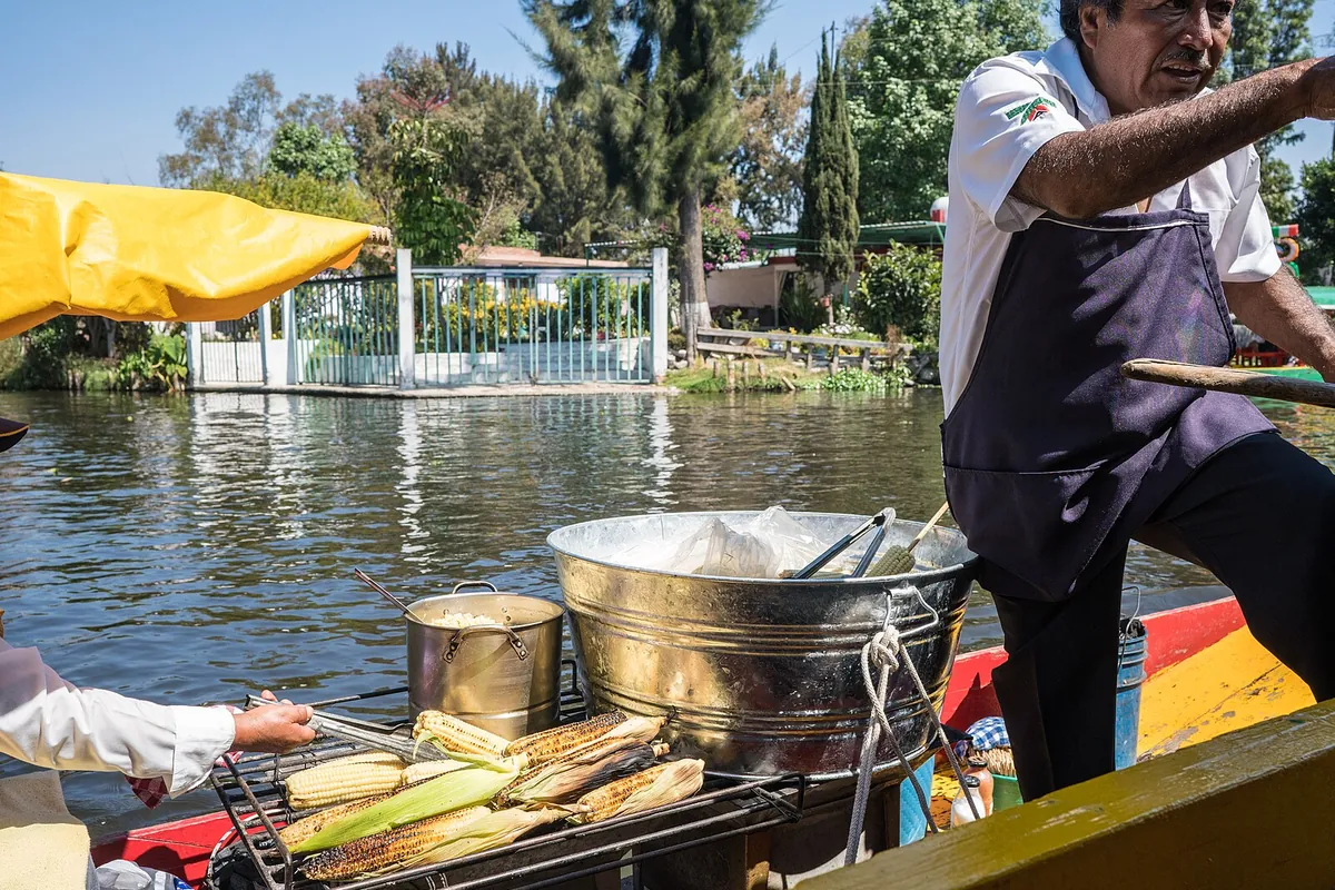 A boatman grilling corn on a charcoal grill on a small boat in a Xochimilco canal, with a customer on another boat reaching over to pay
