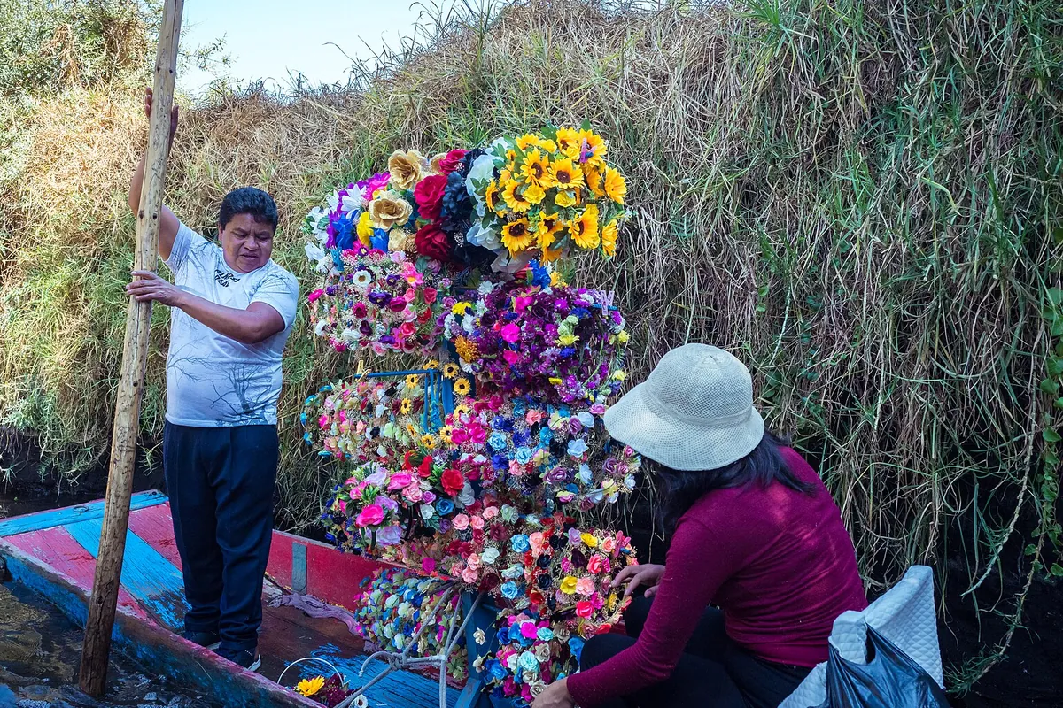 A vendor standing at the stern of a boat piled high with bunches of artificial flowers and flower crowns in a Xochimilco canal